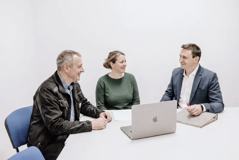Stefan Franz im blauen Sakko rechts an einem Besprechungstisch, im Gespräch mit zwei weiteren Personen. Auf dem Tisch ein Laptop und Unterlagen.